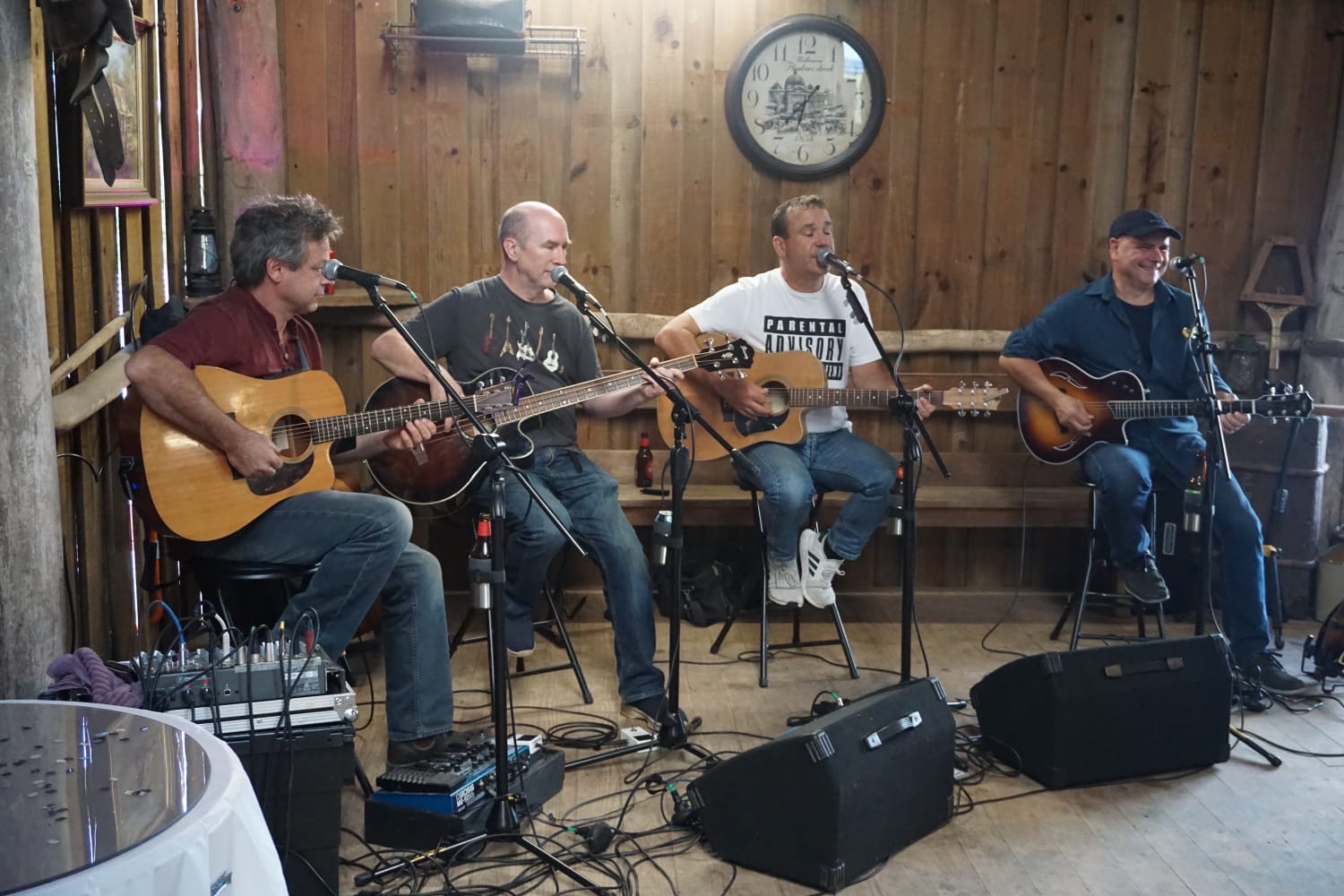 Peter Thompson, Pete Byrne, Jay Harvey and Ron Wenzler playing acoustic guitars and bass on stools in a timber barn